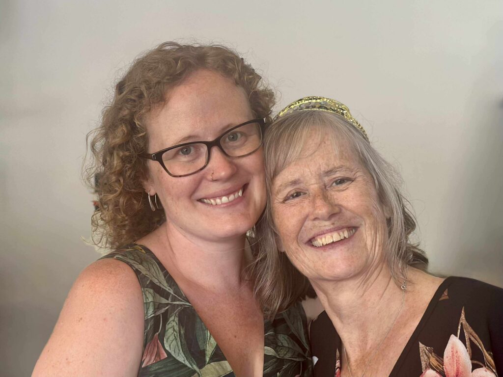 Close-up selfie of two women smiling, one with glasses and curly hair, the other with a gold headband and light hair, hugging together.
