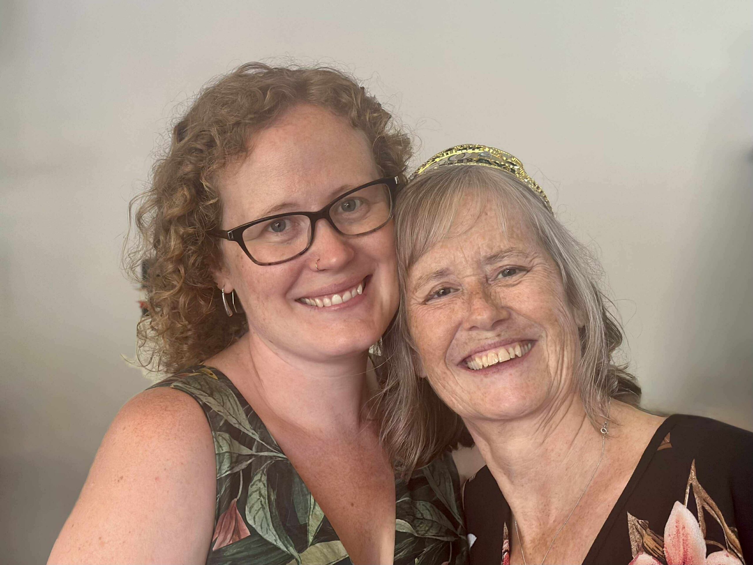 Close-up selfie of two women smiling, one with glasses and curly hair, the other with a gold headband and light hair, hugging together.