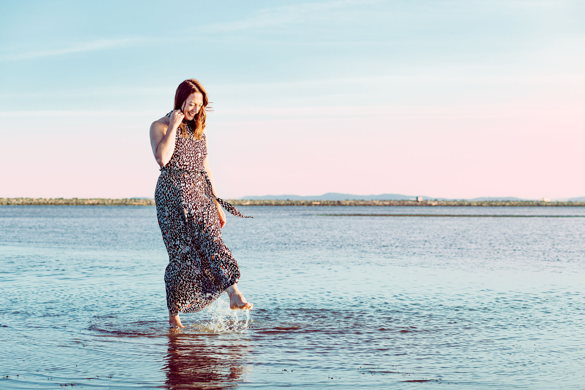 Woman in a patterned dress wading in shallow beach water, smiling as small waves splash around her.