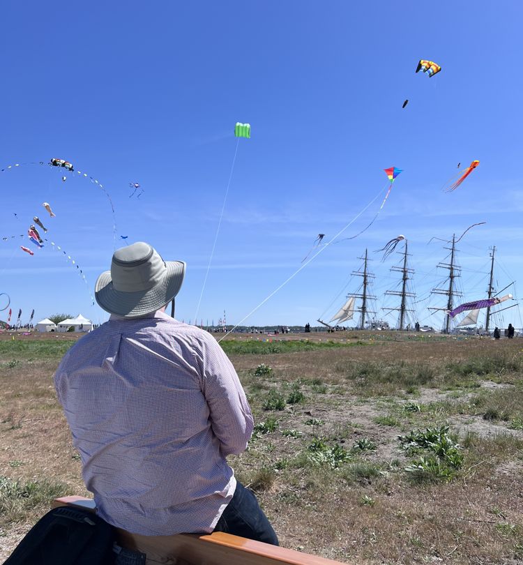 Man with a wide-brimmed hat sits on a bench, watching colorful kites fly in a blue sky near tall ships in the harbor.